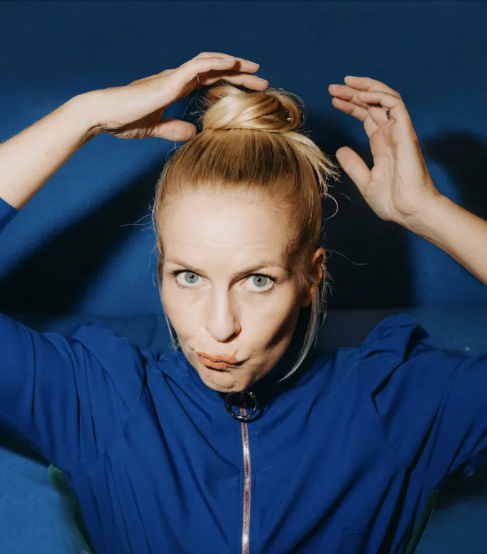 Alexandra von Lingen richtet sich den Dutt auf dem Kopf. Sie trägt ein blaues Oberteil vor einer blauen Wand und verzieht das Gesicht humorvoll
